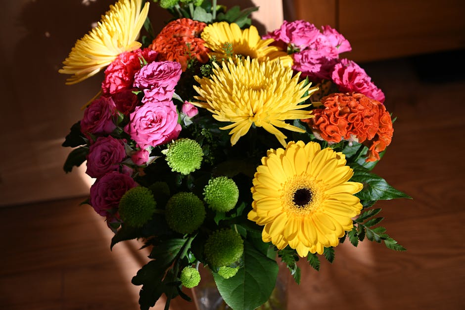 A vibrant floral bouquet arranged in a clear glass vase, featuring bright yellow gerbera daisies with dark centers, pink and purple spray roses, orange carnations, and green button mums. The bouquet i