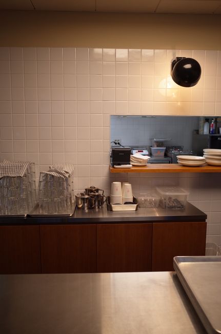 The image depicts a professional kitchen or food preparation area with a white tiled wall and stainless steel countertop. On the left side, there are several glass dessert cups, some covered with mesh