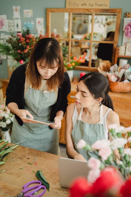 Two women in a florist workshop, wearing light green aprons, are engaged in floral arranging inside a shop decorated with colorful flowers and potted plants. One woman with shoulder-length brown hair 