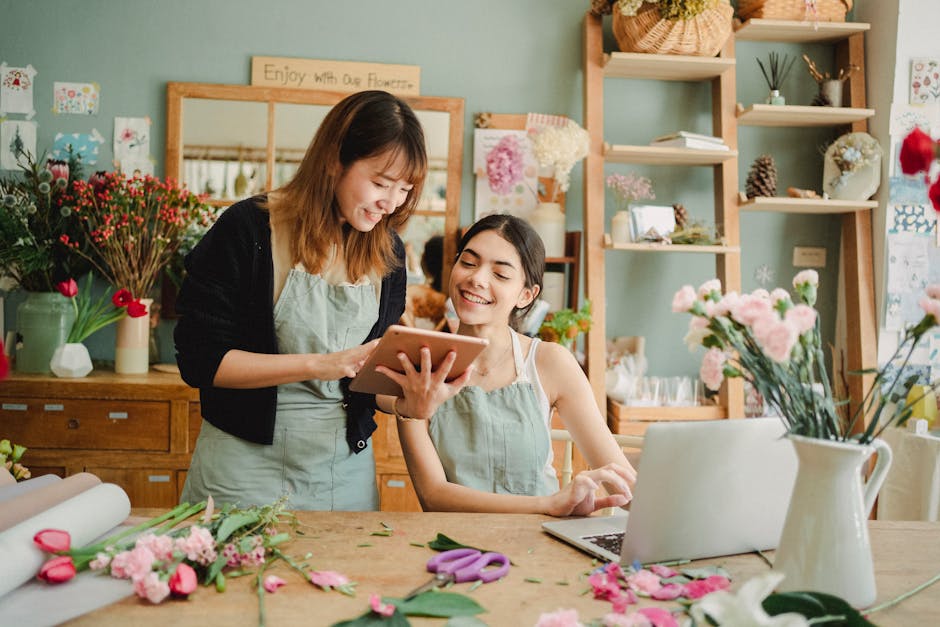 Two women inside a florist shop, one standing and holding a tablet while the other is seated at a wooden worktable, both smiling and looking at the device. The table is decorated with various flowers,