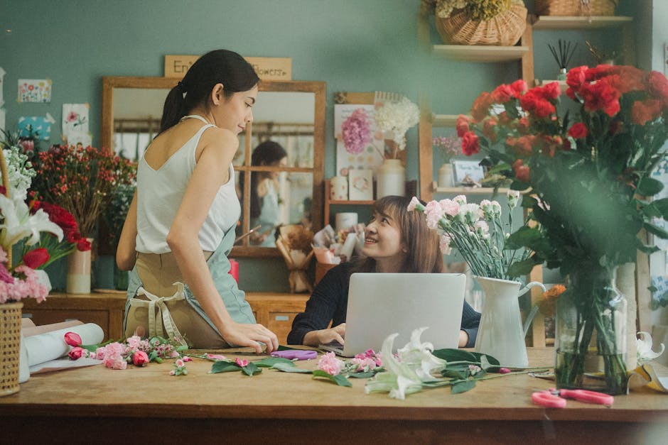 A young woman with long brown hair, sitting at a wooden table inside a florist shop, is smiling up at a woman standing beside her. The woman standing, wearing a white sleeveless top and an apron, is e