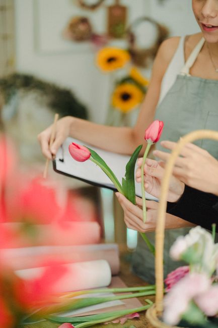 A florist at a workbench in a well-lit shop holds a small bouquet of fresh pink tulips with green stems and leaves, assembling a floral arrangement. The florist is wearing a light-colored apron over a