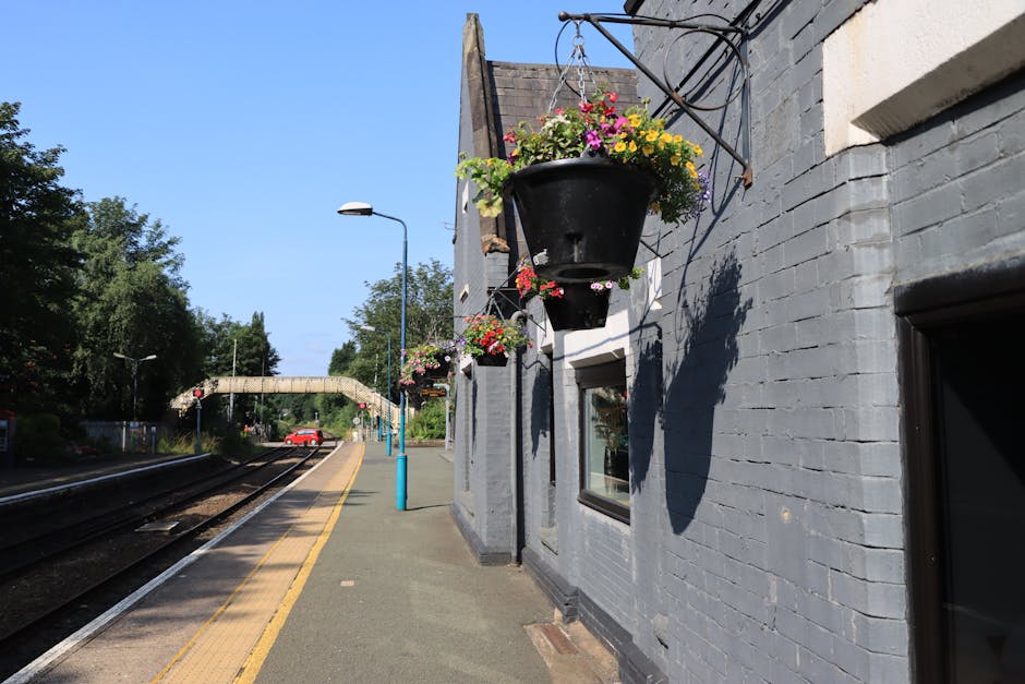 A black hanging flower basket filled with pink, yellow, and purple petunias and geraniums, suspended from a metal bracket on a grey brick wall at Colney Hatch station. The station platform features a 