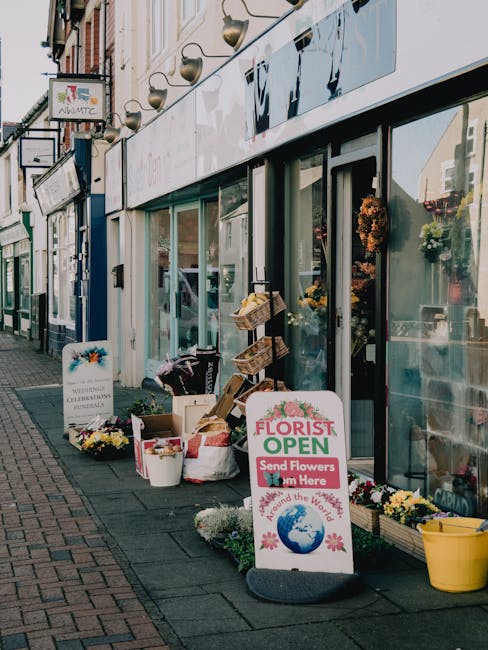 The image shows the exterior of a florist shop with large glass windows displaying a variety of floral arrangements inside. Outside, there are several buckets and baskets filled with freshly cut flowe