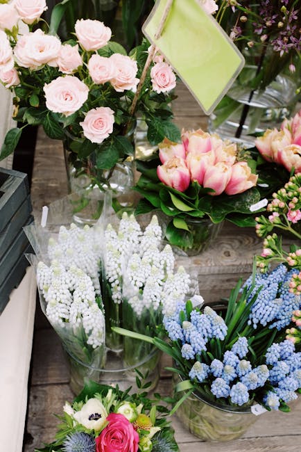 A display of fresh floral arrangements on a rustic wooden table featuring a variety of flowers including light pink roses with lush green foliage arranged in a tall, clear glass vase, pink and yellow 