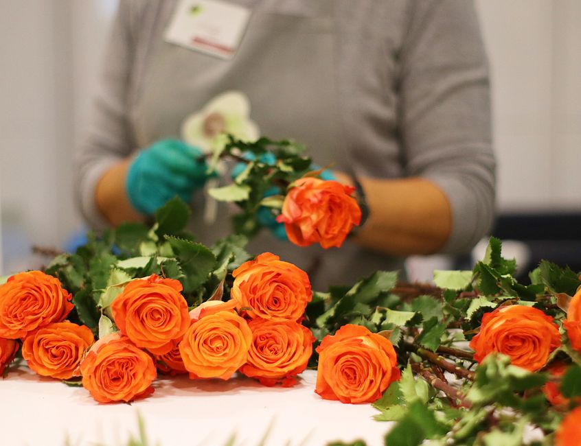 Close-up of a florist from floristcolneyhatch.co.uk assembling a floral arrangement with vibrant orange roses that have densely layered petals and bright green leaves, alongside white orchids with del